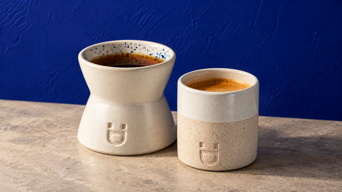 Duas xícaras de cerâmica artesanal DROPIT com acabamento branco e bege, uma com café coado e outra com espresso, sobre bancada de pedra clara e fundo de parede azul texturizada.